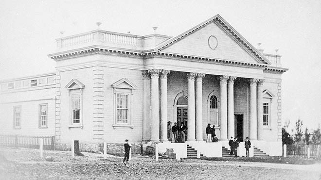 Historic black and white photograph of a neoclassical building with columns, featuring several people gathered on the steps and in front of the entrance.